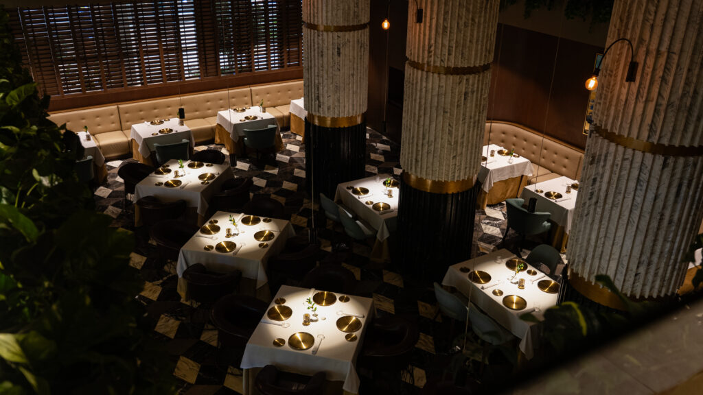 María Dolores by Edgar Núñez Haute Grandeur Global Awards 3 - Petróleo y Energía Upscale dining area with white-tablecloth tables, gold chargers, and booths along a wall, illuminated by warm hanging bulbs.