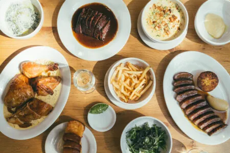 Top-down view of a wooden table with a variety of dishes: roasted chicken, sliced steak in sauce, fries, greens, and side bowls.