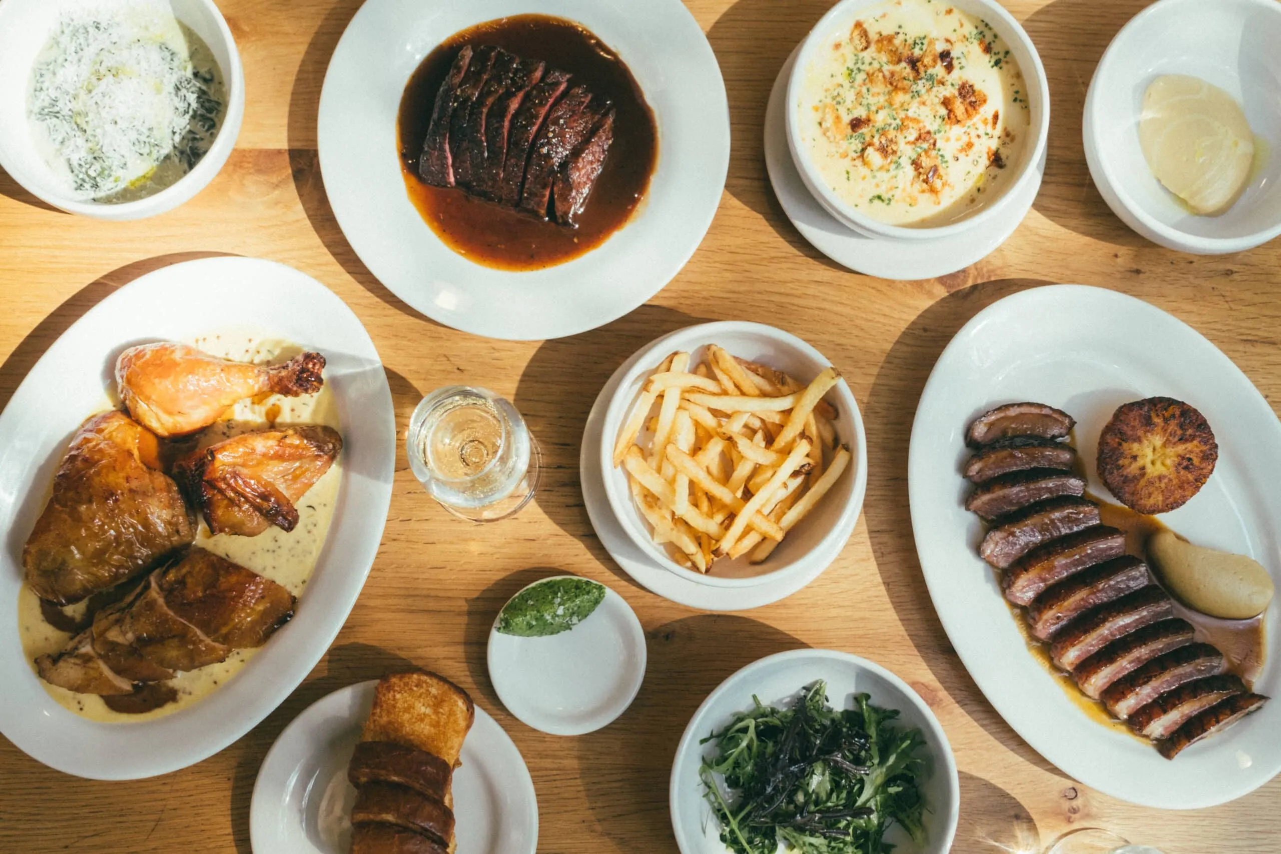 Top-down view of a wooden table with a variety of dishes: roasted chicken, sliced steak in sauce, fries, greens, and side bowls.