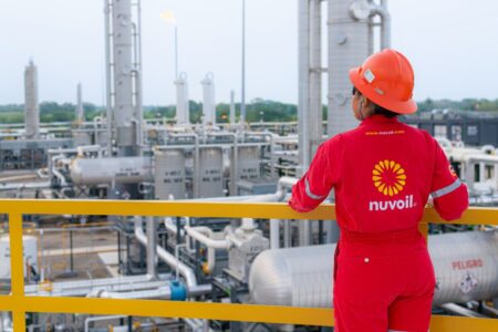 Industrial worker in red protective suit and helmet overlooking a chemical/refinery plant with metal pipes and tanks, seen from behind on a yellow railing.
