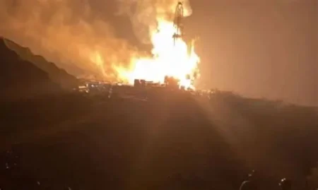 A large fire on a hillside at night, flames rising and illuminating smoke as a pile of debris burns.