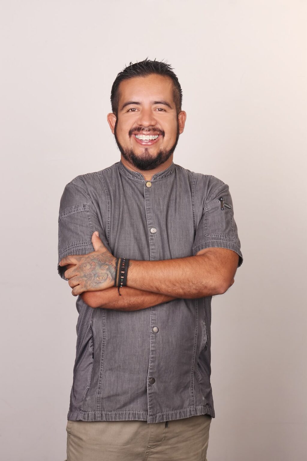 PHOTO-2026-07-07-14-30-08 - Petróleo y Energía Man standing with arms crossed, smiling, wearing a gray short-sleeve button-up shirt; tattoos on his left forearm visible.