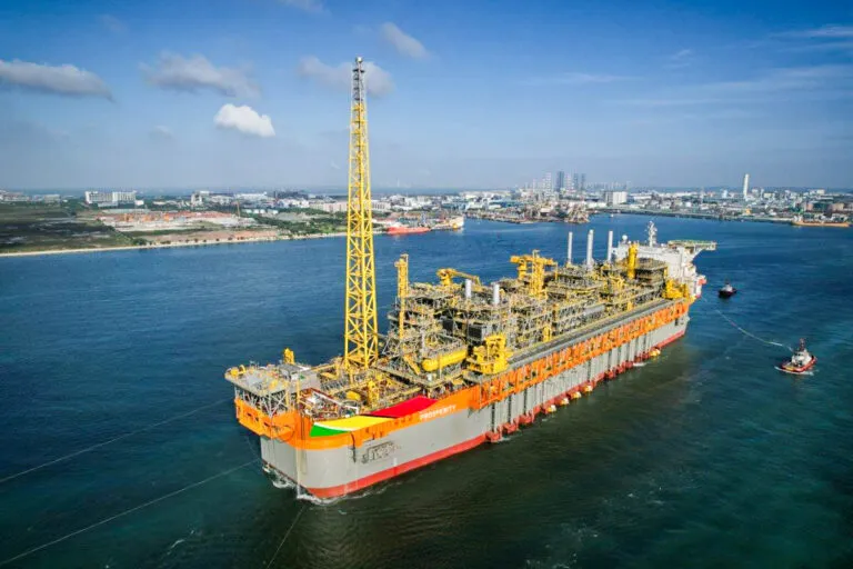 Large orange offshore construction vessel with yellow cranes floating in a harbor, city skyline in the distance under a blue sky