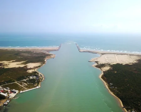 Aerial view of a coastal channel with parallel breakwaters opening to the sea, flanked by sandy shores and sparse vegetation on both sides.