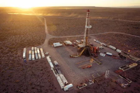 Oil drilling rig in a desolate desert oil field, with trailers and equipment around the base at sunset or golden hour.