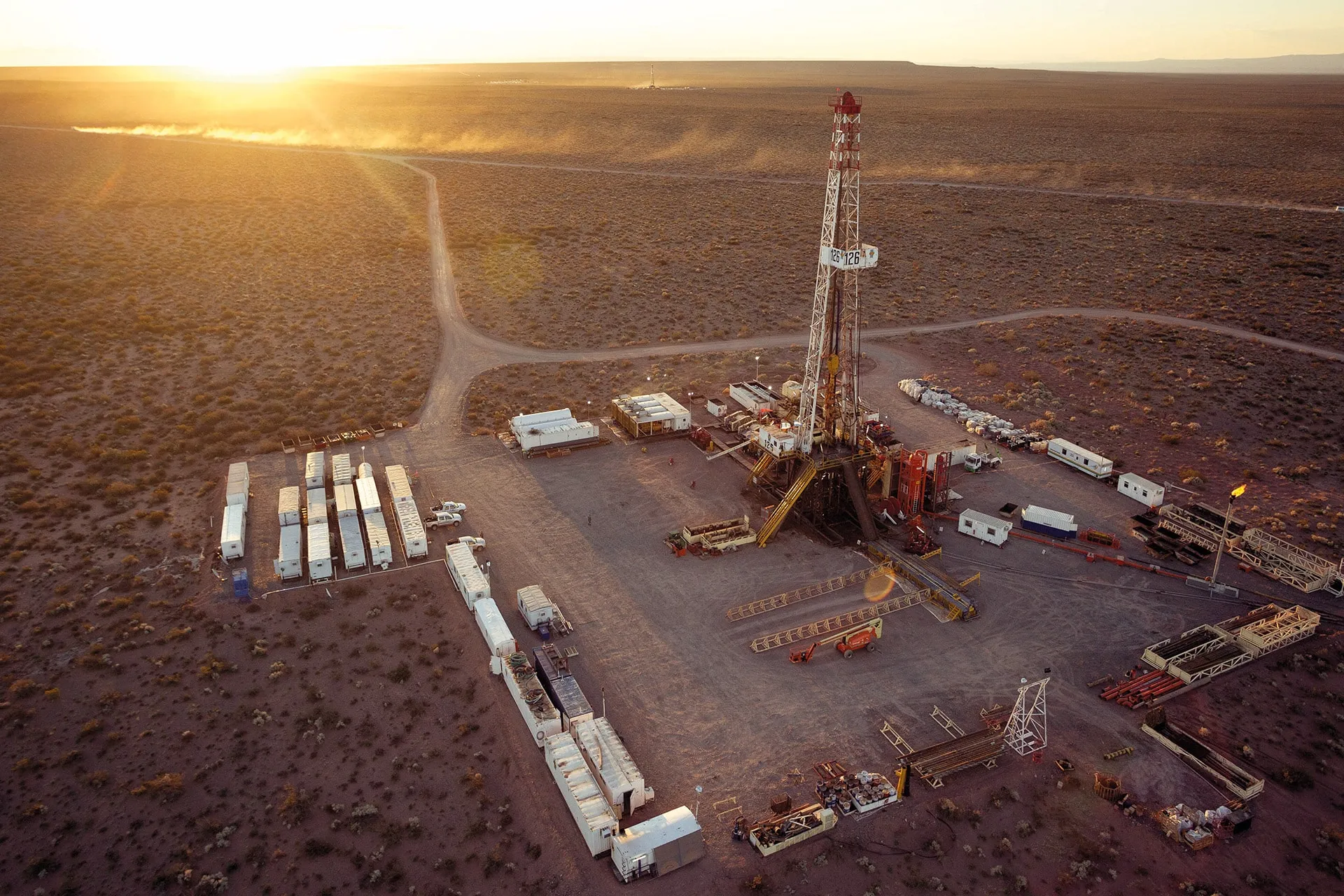 Oil drilling rig in a desolate desert oil field, with trailers and equipment around the base at sunset or golden hour.