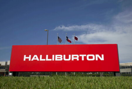 Red Halliburton sign with three flagpoles (US, Texas, red flag) against a blue sky.