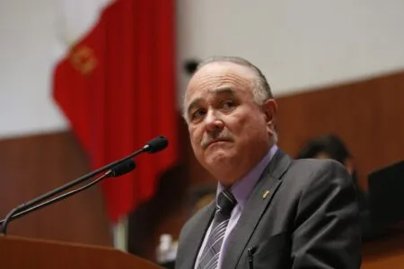 Older man in a gray suit giving a speech at a podium, microphone in front, red flag in background.