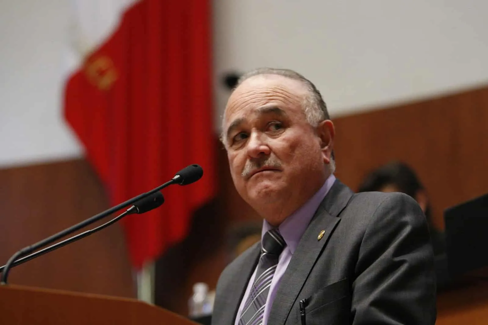 Older man in a gray suit giving a speech at a podium, microphone in front, red flag in background.