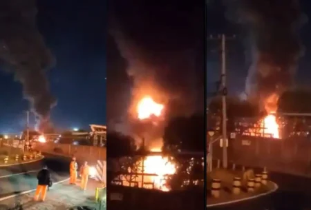 Nighttime industrial fire with tall flames and thick smoke; workers in orange suits stand near a barricade nearby.