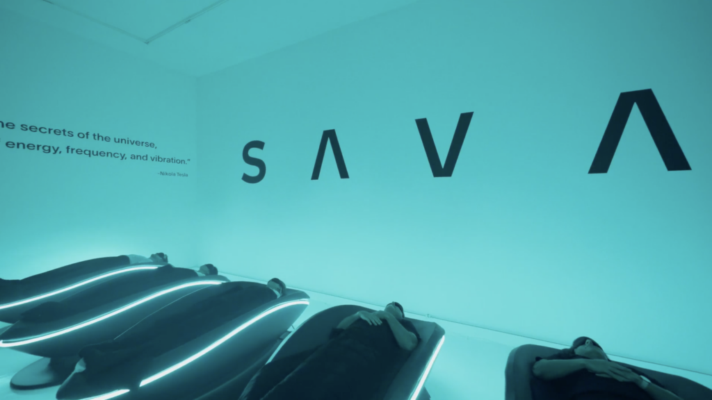 SAVA X L´Ermitage Beverly Hills 2 - Petróleo y Energía Blue-walled room with large'SAVA' letters and a Nikola Tesla quote; visitors rest in curved, neon-lit pods with a blue glow.