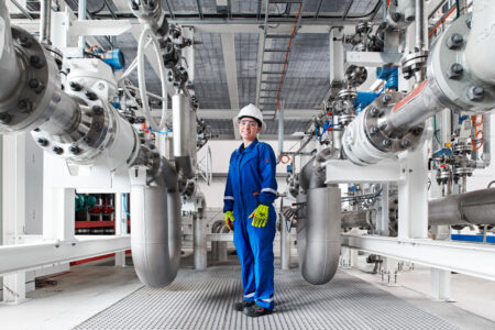Maintenance worker in blue coveralls and a white hard hat stands among large stainless steel pipes and valves in an industrial facility, safety gloves on.