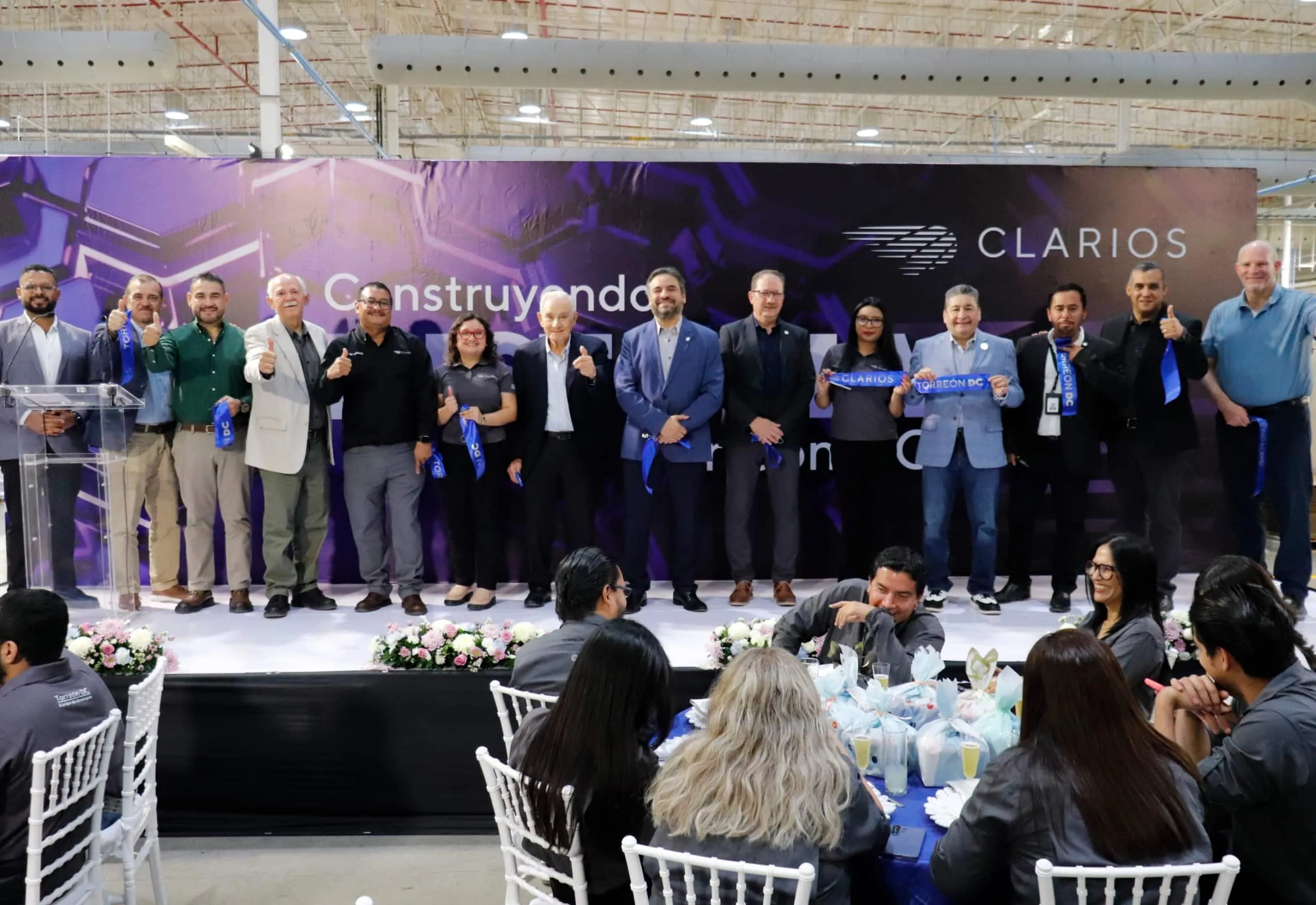 Group of professionals on stage posing with thumbs up at a CLARIOS event, purple backdrop behind them.