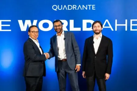 Two men in suits shake hands at a corporate event, with a third man smiling beside them in front of a blue Quadrante banner.