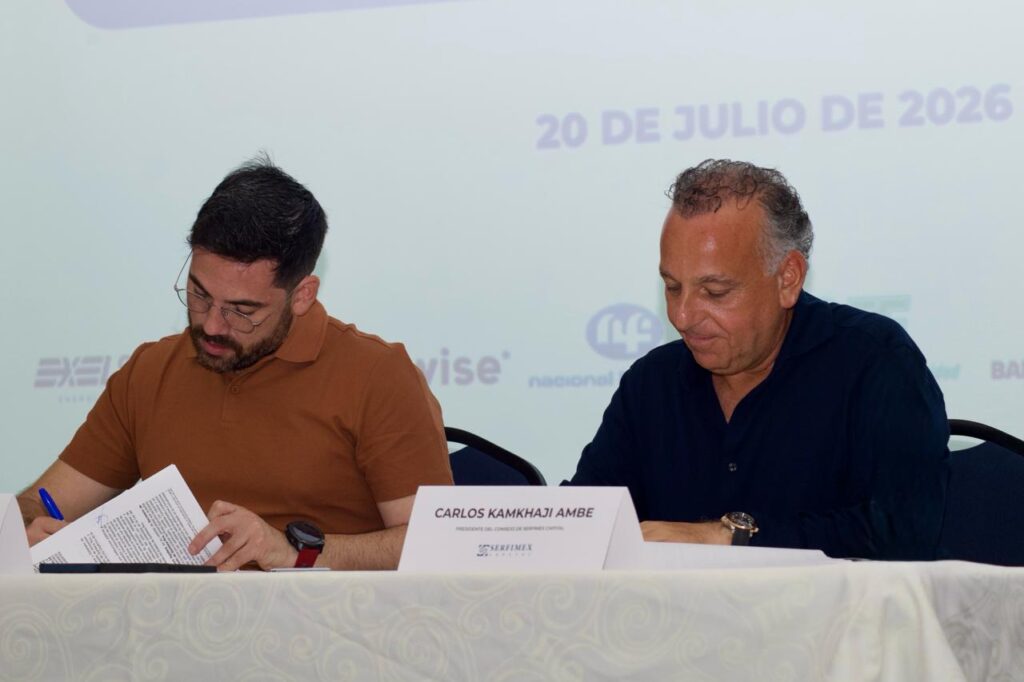 WhatsApp Image 2026-07-21 at 95048 AM - Petróleo y Energía Two men sit at a table signing documents at a formal signing event; one in a tan polo, the other in a dark blue shirt, with a name card visible on the table.