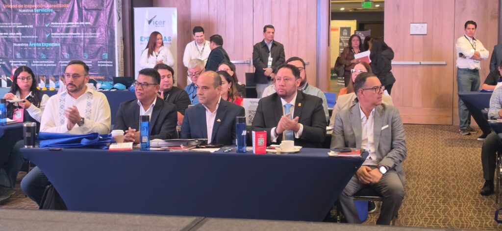 WhatsApp Image 2026-07-30 at 92240 AM 1 - Petróleo y Energía Panel of five men in suits seated at a blue-draped table during a conference, with an attentive audience behind them.