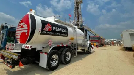 PEMEX tanker truck parked at an oil field, with orange-suited workers and drilling equipment in the background