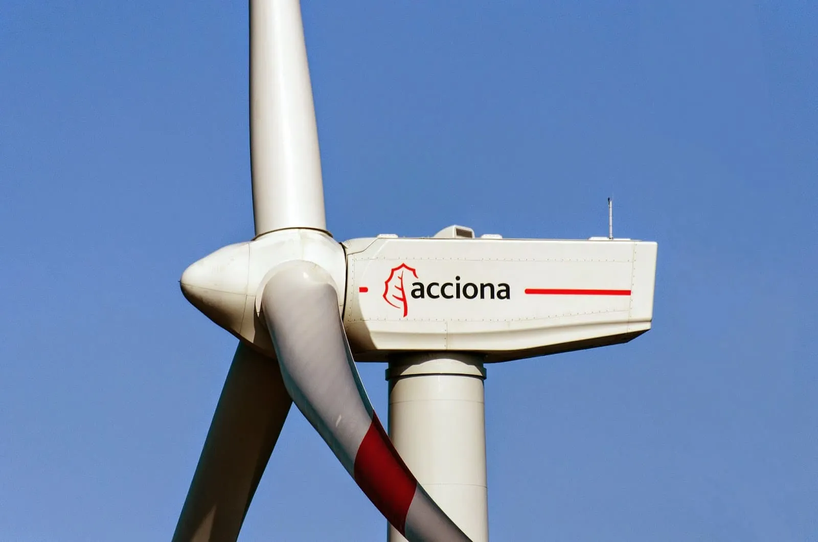 Wind turbine rotor and nacelle with the Acciona logo against a clear blue sky, close-up view of the blades and tower.