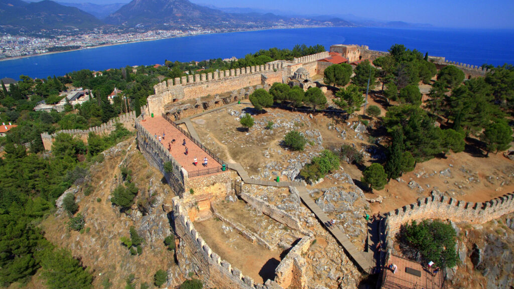 alanya-castle-4 - Petróleo y Energía A medieval fortress with stone walls on a hillside, overlooking a blue sea and coastline.