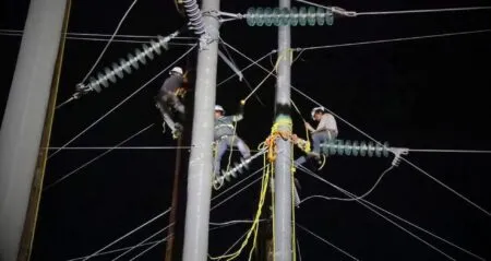 Nighttime scene of utility workers on a tall power pole making repairs to overhead lines, wearing hard hats and safety gear with ropes and insulators present