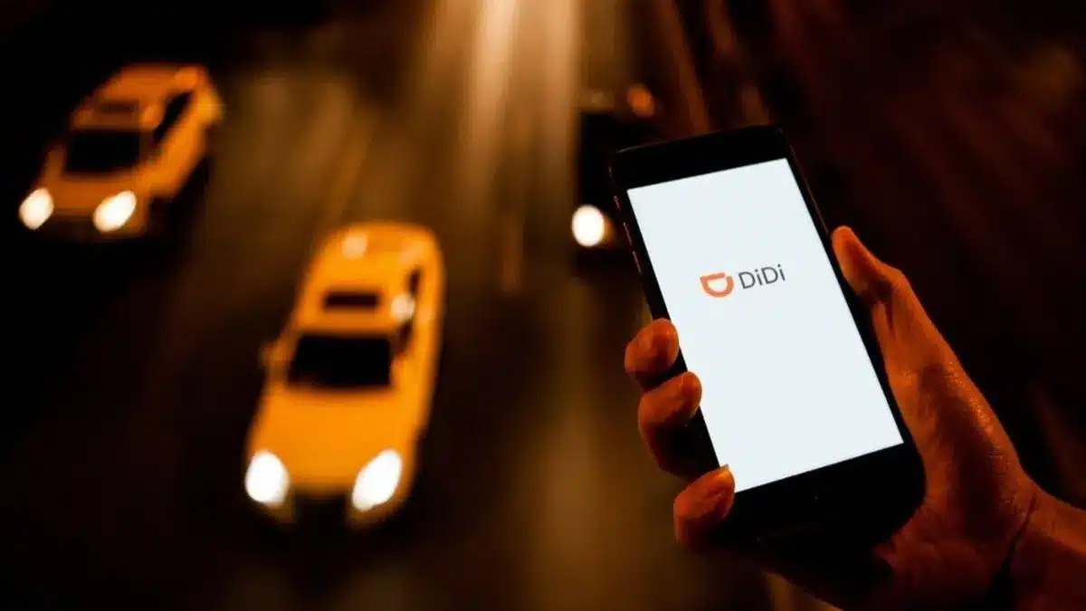 Person holding a phone displaying the DiDi app logo as yellow taxis drive by on a nighttime city street.