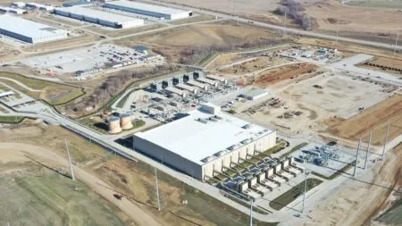 Aerial view of an industrial complex with a large white warehouse, several processing units, and storage tanks surrounded by cleared construction areas and roads.