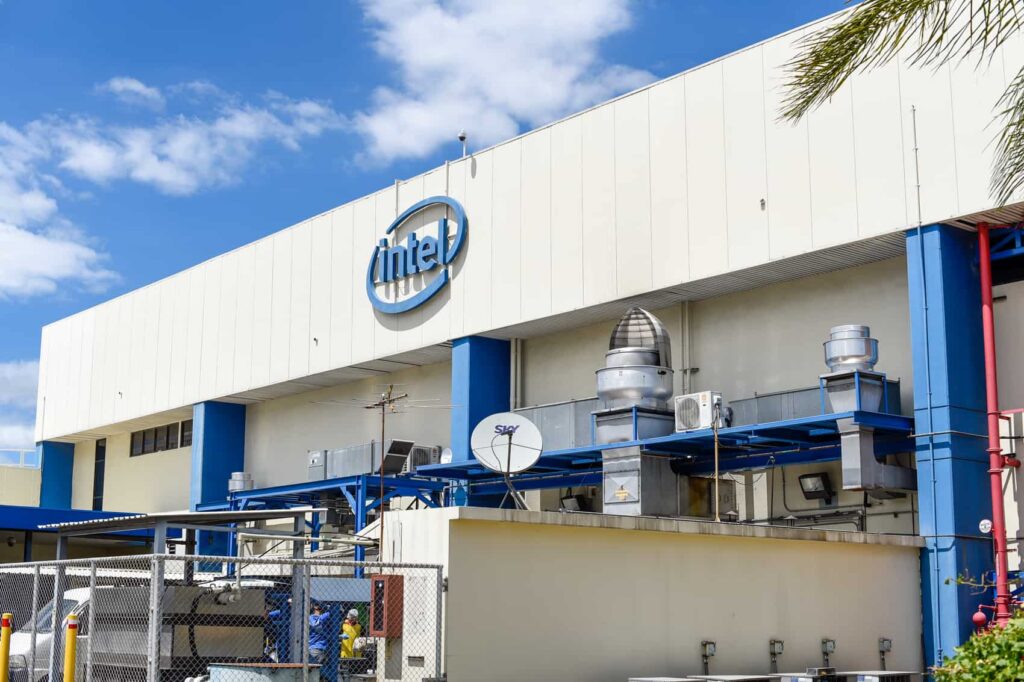 Exterior view of a white industrial building with the Intel logo on the facade and blue structural accents under a blue sky.