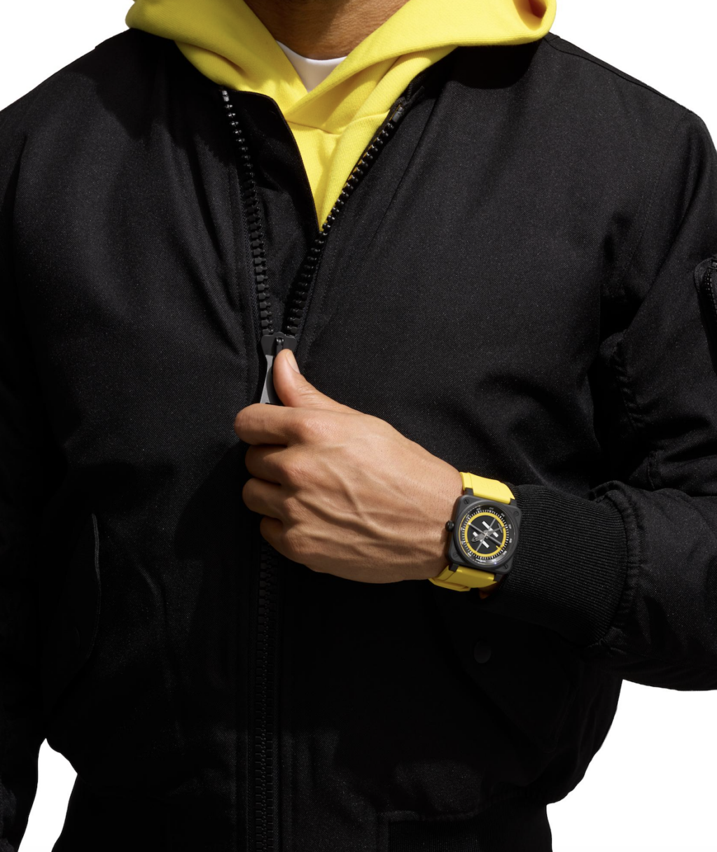 helipad 4 - Petróleo y Energía Person in a black zip-up jacket over a yellow hoodie, pulling the zipper open near the chest; yellow watch on left wrist