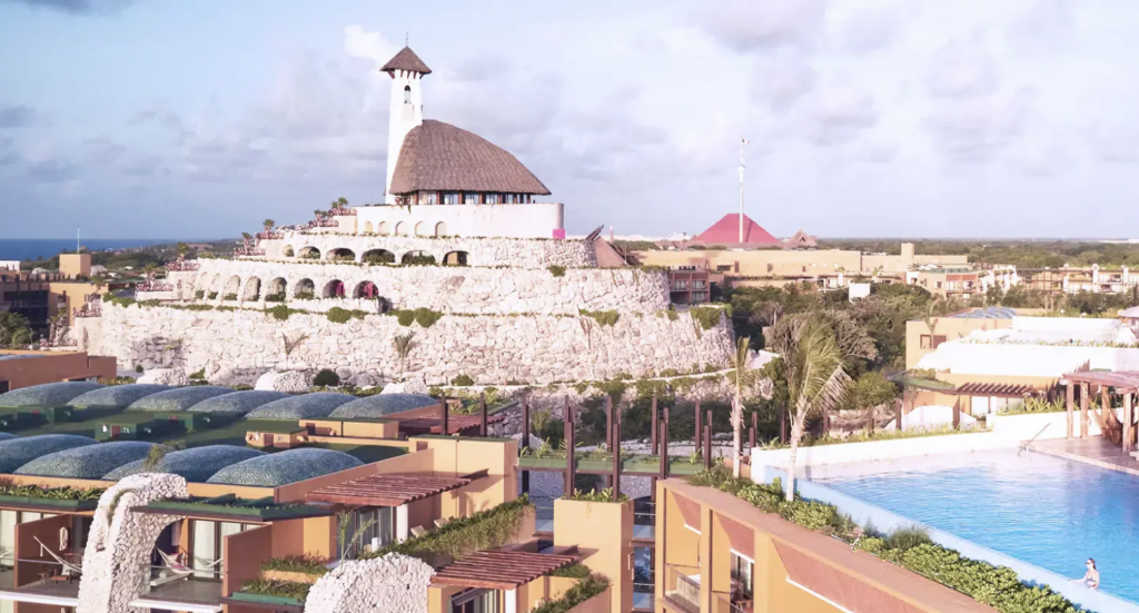 hotel xcaret arte 2 - Petróleo y Energía Resort complex with a tiered white stone fortress, arched openings, and a thatched-roof building overlooking a pool area.