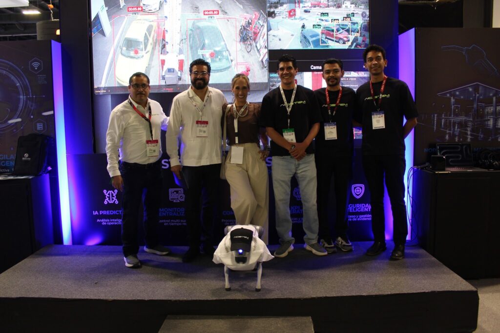 image - Petróleo y Energía Five people with badges posing on a stage at a tech expo, in front of a large video wall displaying cars; a white robotic prop sits on the floor in front.