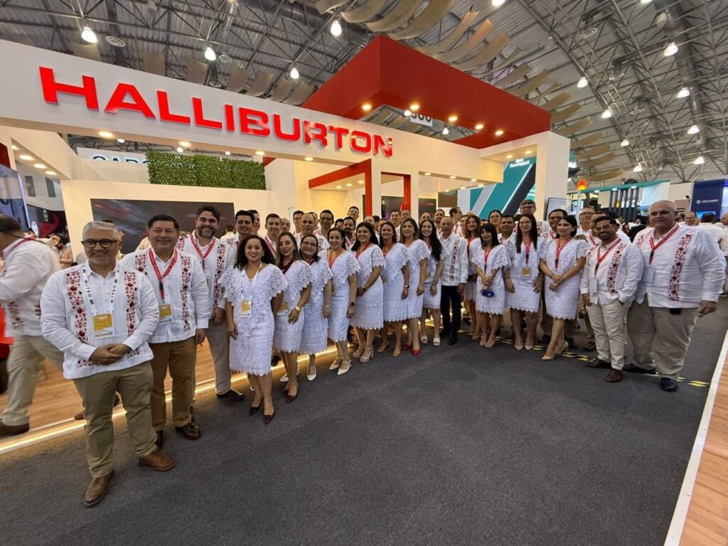 image - Petróleo y Energía Group of people in white embroidered shirts posing for a group photo at a Halliburton booth in a trade show hall, wearing red lanyards and name badges.