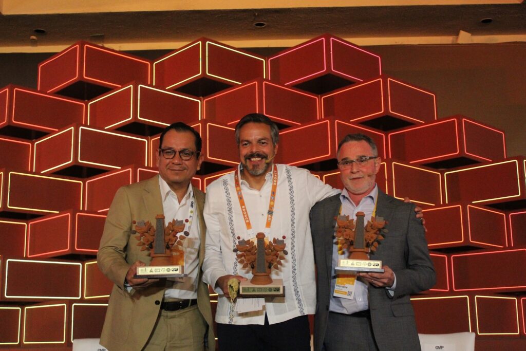 image - Petróleo y Energía Three men in suits pose with wooden trophies on a stage with a red neon cube backdrop, smiling for the photo.