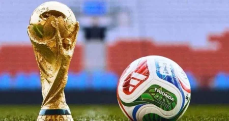 Golden trophy standing beside a colorful soccer ball on a grass field with stadium seating blurred in the background