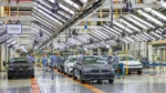 Automotive assembly plant with multiple cars on a moving bright orange conveyor in a spacious, brightly lit aisle; workers in blue uniforms inspect vehicles under overhead machinery.