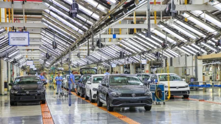 Automotive assembly plant with multiple cars on a moving bright orange conveyor in a spacious, brightly lit aisle; workers in blue uniforms inspect vehicles under overhead machinery.