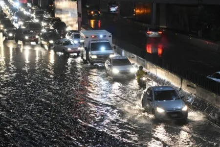 Nighttime street flooded with moving cars, a motorcycle, and an ambulance amid heavy traffic with glowing reflections on the water surface.
