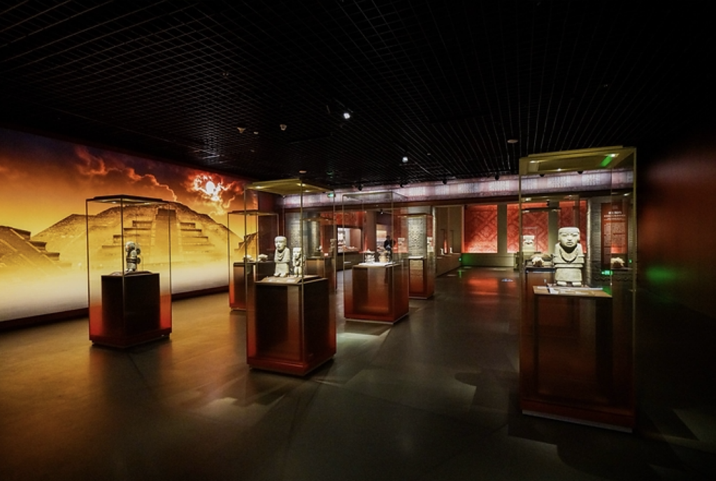 museo shangai 3 - Petróleo y Energía Museum gallery with glass display cases showcasing small statues; warm pyramid mural along the wall in the background.