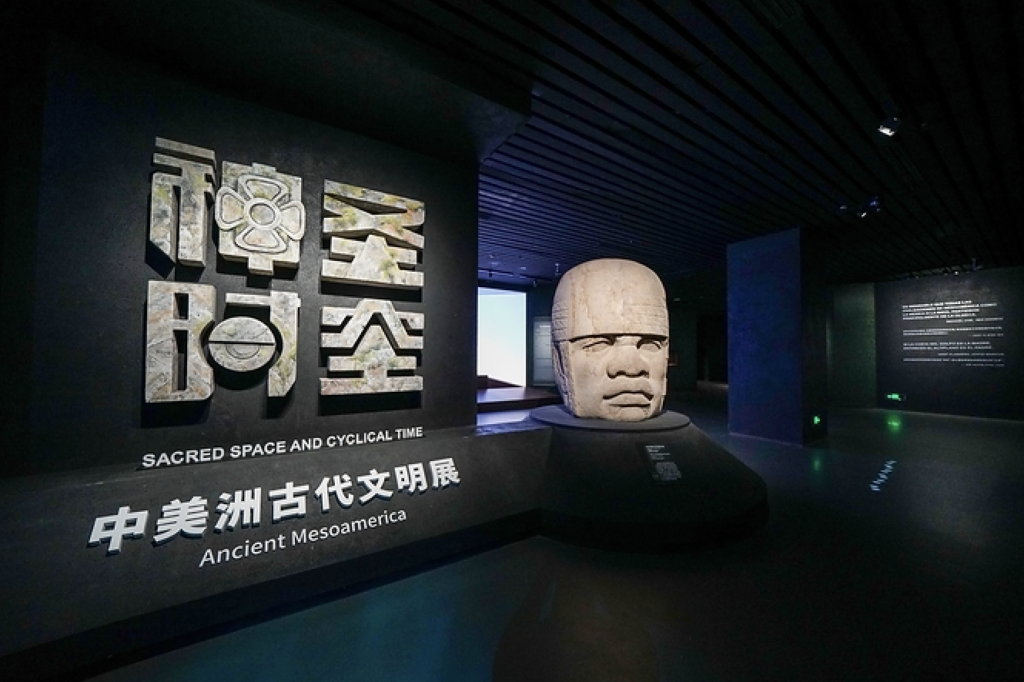 museo snagai 2 - Petróleo y Energía Museum exhibit with a large ancient stone head sculpture and bold signage reading'SACRED SPACE AND CYCLICAL TIME' and 'Ancient Mesoamerica' in English and Chinese flags the display area.