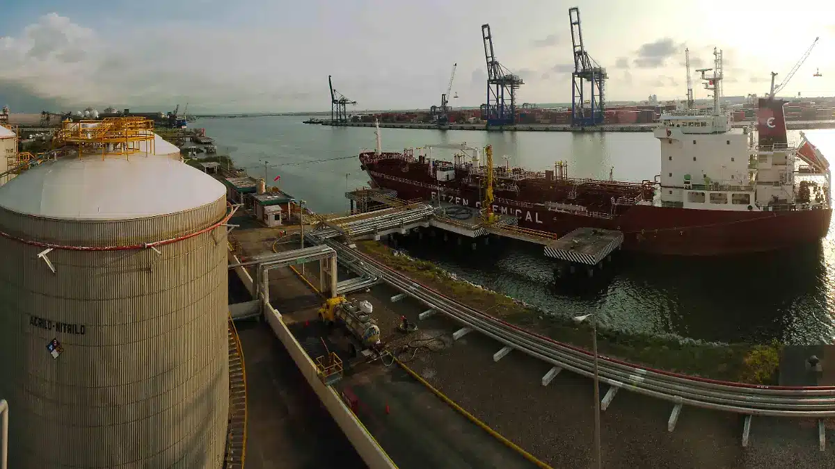 Industrial port scene with a large storage tank in the foreground, a red chemical tanker docked at the quay, and blue gantry cranes across the water.