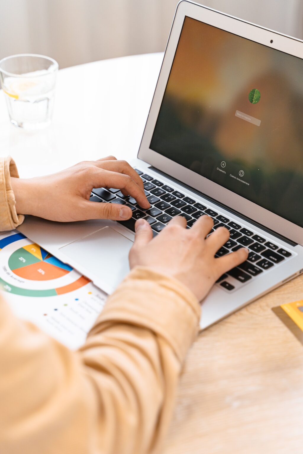 pexels-ai25studio-8374254 - Petróleo y Energía Person typing on a silver laptop at a desk, with a glass of water and a pie chart in view.