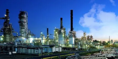Industrial refinery complex at night with tall distillation columns and extensive piping, illuminated by blue-toned lights.