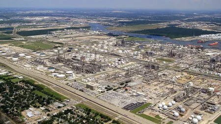 Aerial view of a large oil refinery with many storage tanks, towers, and pipelines by a river.