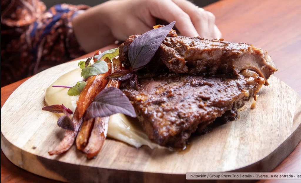 resta mestizo - Petróleo y Energía Succulent glazed roasted meat with a crispy crust served on a wooden board, garnished with colorful herbs and carrot ribbons.