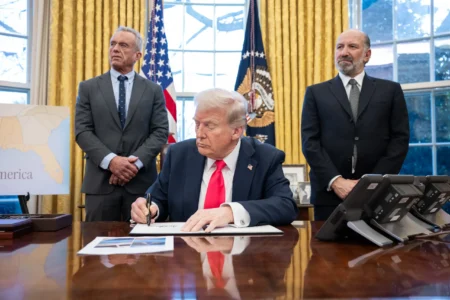 President Donald Trump signs a document at a desk, flanked by two suited men in a room with gold curtains and American flags behind him.