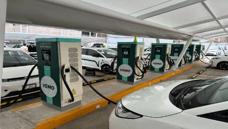 Row of electric car chargers labeled VEMO under a covered canopy, each connected to parked cars in a charging station lot.