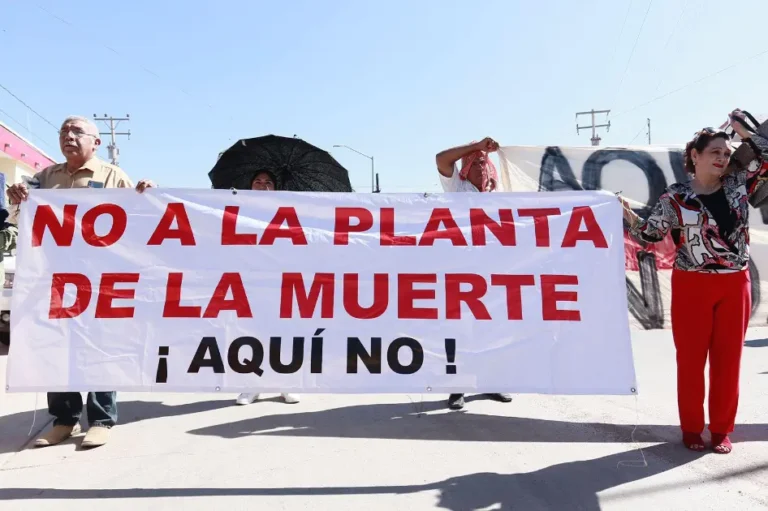 Protesters hold a large white banner reading 'NO A LA PLANTA DE LA MUERTE ¡AQUÍ NO!' in red and black, outdoors on a sunny day.