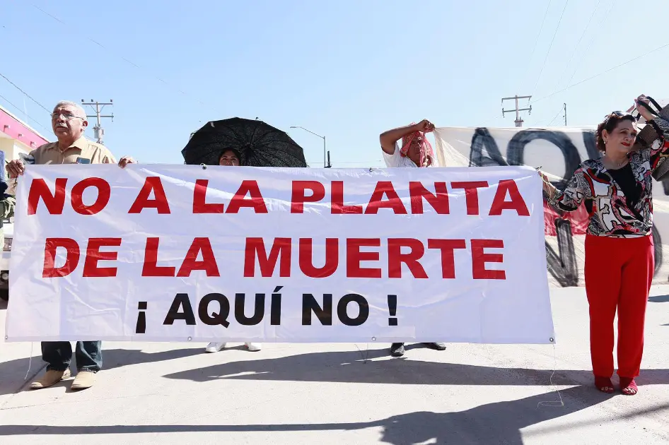 Protesters hold a large white banner reading 'NO A LA PLANTA DE LA MUERTE ¡AQUÍ NO!' in red and black, outdoors on a sunny day.