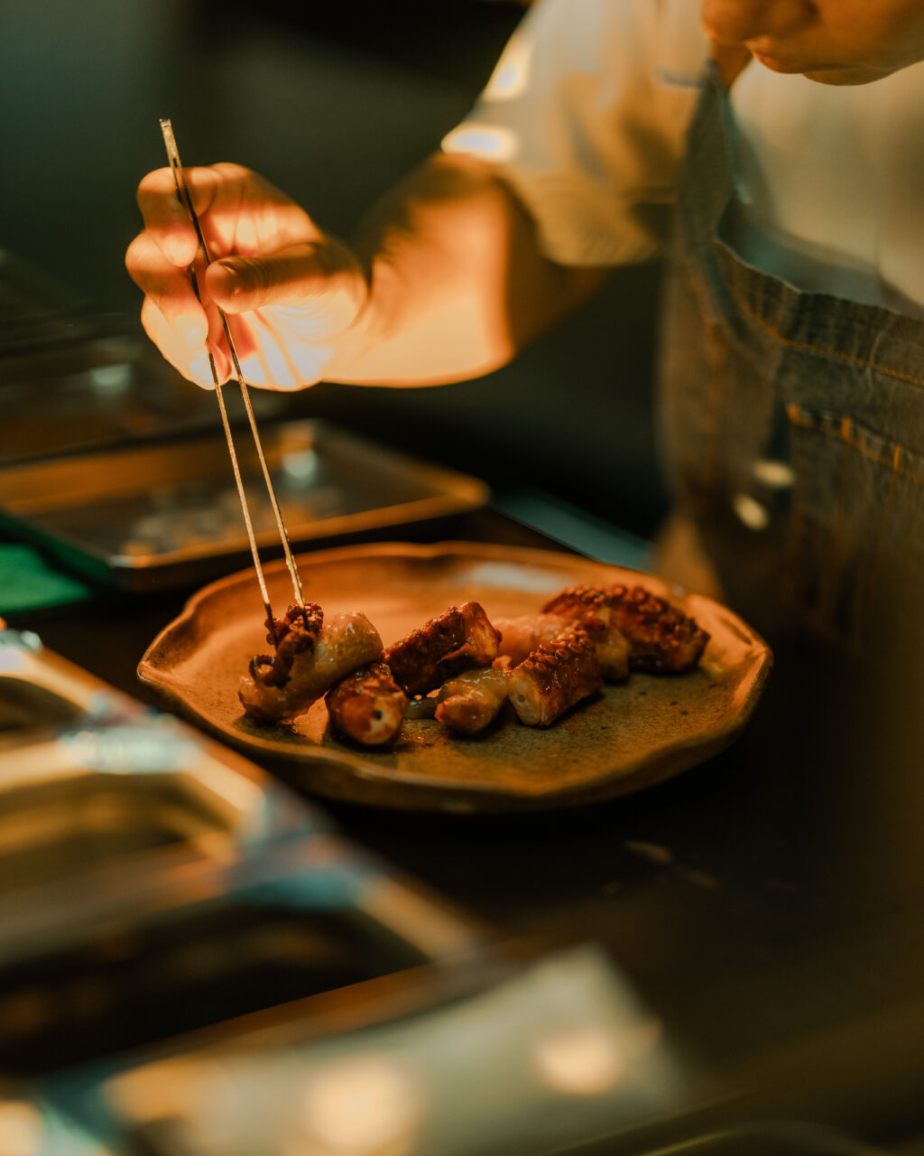 0I5A3081 - Petróleo y Energía Chef arranging skewered meat bites on a plate under warm kitchen lighting with tongs in hand