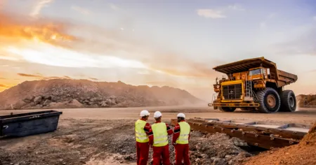 Three workers in high-visibility vests and hard hats stand together at a mining site, with a large dump truck in the background at sunset.