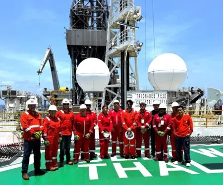 Group of workers in red coveralls and white hard hats standing on a helipad in front of an offshore drilling rig.